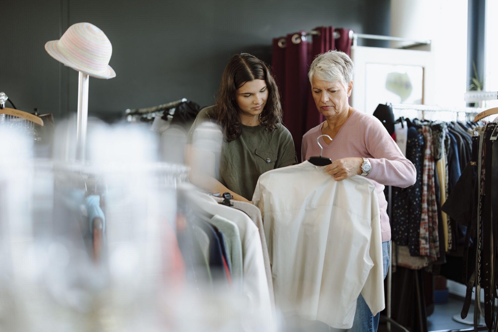 Eine &auml;ltere Dame mit grauem Haar steht in einem Secondhandladen und h&auml;lt eine wei&szlig;e Bluse in der Hand. Neben ihr steht ein junges M&auml;dchen mit lockigem Haar und einem olivgr&uuml;nen T-Shirt, das ebenfalls die Bluse interessiert betrachtet.