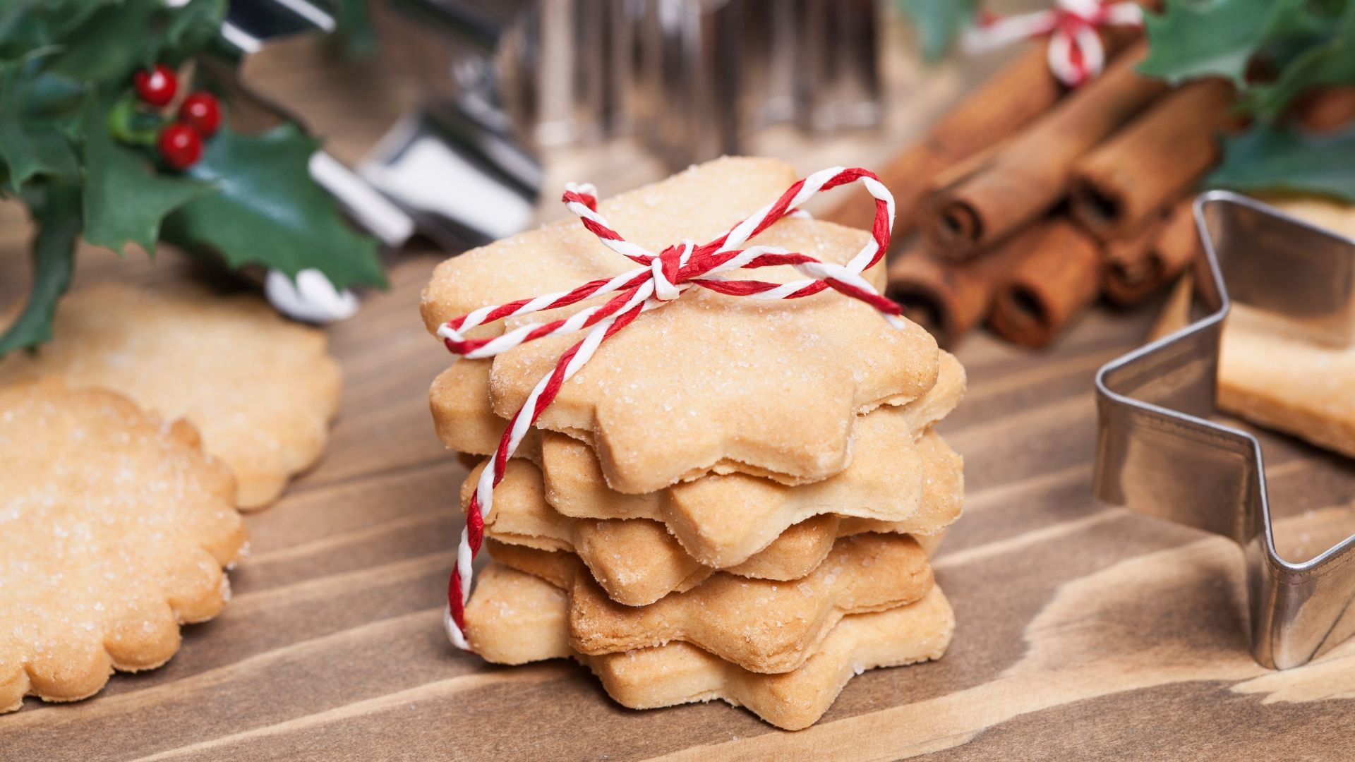 Auf dem Bild sind ein Stampel Kekse in Sternform zusehen, die mit einer rot-wei&szlig;en Schnurr zusammengebunden sind. Im Hintergrund sind Keksausstecher sowie Zimtstanden zusehen.